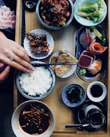 A wooden tray holds a variety of Japanese dishes including a bowl of steamed white rice, a small serving of sashimi with slices of salmon and tuna, a dish of cooked eel with sauce, a vibrant green salad, and several small bowls containing various appetizers such as seaweed, miso soup with mushrooms, and a dipping sauce. A person’s hand with a ring is placing chopsticks beside the rice bowl, suggesting preparation to eat.