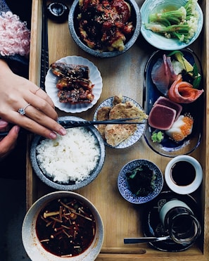 A wooden tray holds a variety of Japanese dishes including a bowl of steamed white rice, a small serving of sashimi with slices of salmon and tuna, a dish of cooked eel with sauce, a vibrant green salad, and several small bowls containing various appetizers such as seaweed, miso soup with mushrooms, and a dipping sauce. A person’s hand with a ring is placing chopsticks beside the rice bowl, suggesting preparation to eat.