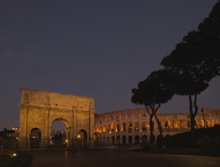 A historical archway stands prominently illuminated against the night sky, accompanied by a large ancient amphitheater partially lit with warm tones. The scene is framed by dark silhouettes of trees.