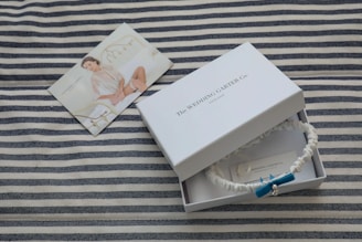 A striped fabric serves as the background for a white box labeled 'The Wedding Garter Co. England.' Inside the box is a white garter adorned with a blue bow and a small pearl. Beside the box, a photograph of a woman wearing bridal lingerie is displayed, resting on the fabric.