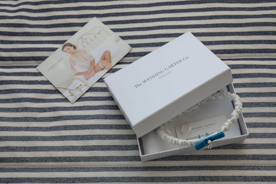 A striped fabric serves as the background for a white box labeled 'The Wedding Garter Co. England.' Inside the box is a white garter adorned with a blue bow and a small pearl. Beside the box, a photograph of a woman wearing bridal lingerie is displayed, resting on the fabric.