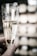 A hand holds a champagne flute filled with sparkling wine, with bubbles visible in the liquid. The background is softly blurred, showcasing a wine rack filled with bottles.