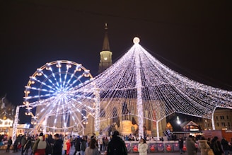 A festive scene featuring a large Ferris wheel adorned with bright white and blue lights set against a backdrop of a church with a tall steeple. A canopy of twinkling string lights forms a conical tree-like shape, illuminating the busy square filled with people. Small stalls line the area, contributing to a lively holiday market atmosphere.