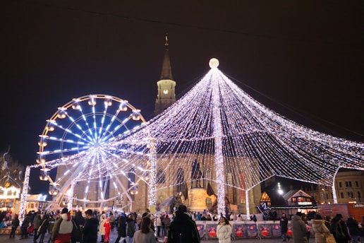 A festive scene featuring a large Ferris wheel adorned with bright white and blue lights set against a backdrop of a church with a tall steeple. A canopy of twinkling string lights forms a conical tree-like shape, illuminating the busy square filled with people. Small stalls line the area, contributing to a lively holiday market atmosphere.