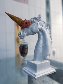 A white unicorn head sculpture is positioned on a pedestal with an ice cream cone placed over its forehead, resembling a horn. The cone is topped with brown ice cream. In the background, there is a blurred image of a jar filled with colorful candies.