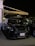 A black Mini Cooper is parked under an awning at night. The car is in a shaded area with ambient lighting highlighting its sleek design. A license plate with Arabic text is visible, and other vehicles are parked nearby.