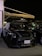 A black Mini Cooper is parked under an awning at night. The car is in a shaded area with ambient lighting highlighting its sleek design. A license plate with Arabic text is visible, and other vehicles are parked nearby.