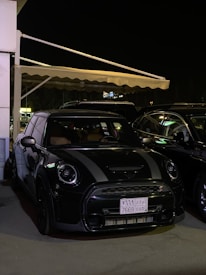 A black Mini Cooper is parked under an awning at night. The car is in a shaded area with ambient lighting highlighting its sleek design. A license plate with Arabic text is visible, and other vehicles are parked nearby.