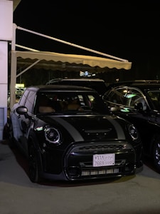 A black Mini Cooper is parked under an awning at night. The car is in a shaded area with ambient lighting highlighting its sleek design. A license plate with Arabic text is visible, and other vehicles are parked nearby.