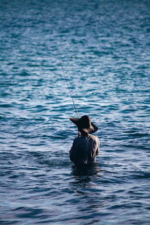 A person wearing a wide-brimmed hat stands waist-deep in the water while holding a fishing pole. The scene captures the tranquility of open water, with gentle waves creating a textured pattern across the surface.