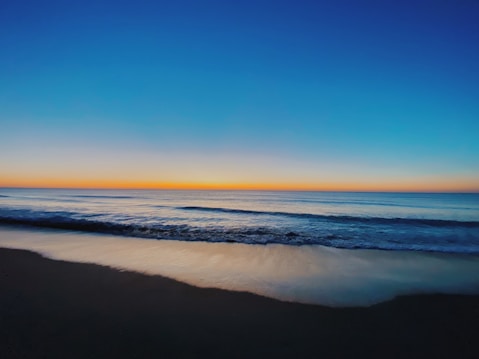 A serene ocean scene with gentle waves lapping against a sandy shore. The sky transitions from a deep blue at the top to a warm orange near the horizon, indicating either sunrise or sunset.