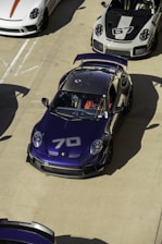 A collection of sports cars parked on a concrete surface, prominently featuring a dark blue car with the number 70 on its hood. Surrounding it are other high-performance cars in white and gray colors, including one with the number 67 on the hood. The cars have aerodynamic features and racing decals, suggesting they are designed for speed and performance.