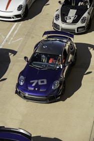 A collection of sports cars parked on a concrete surface, prominently featuring a dark blue car with the number 70 on its hood. Surrounding it are other high-performance cars in white and gray colors, including one with the number 67 on the hood. The cars have aerodynamic features and racing decals, suggesting they are designed for speed and performance.