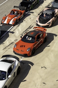 A lineup of high-performance sports cars is positioned on a racetrack. The cars are of various models and colors, including a prominent orange car with the number 21, an orange open-top car with the number 617, a sleek black car, and a white car. Drivers in racing gear are seated in some of the vehicles, preparing for a potential race. The scene conveys a sense of competition and speed, set in a sunny environment with shadows cast by the intense sunlight.