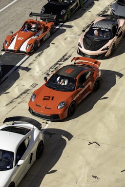 A lineup of high-performance sports cars is positioned on a racetrack. The cars are of various models and colors, including a prominent orange car with the number 21, an orange open-top car with the number 617, a sleek black car, and a white car. Drivers in racing gear are seated in some of the vehicles, preparing for a potential race. The scene conveys a sense of competition and speed, set in a sunny environment with shadows cast by the intense sunlight.