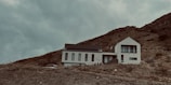 A modern house with a minimalist design is situated on a hillside. The building has a geometric, angular shape with large windows, and it is surrounded by a barren, rocky landscape. The sky is overcast, adding a moody and dramatic atmosphere.