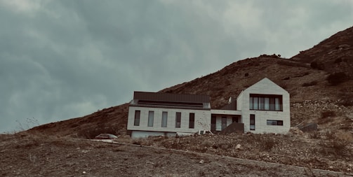 A modern house with a minimalist design is situated on a hillside. The building has a geometric, angular shape with large windows, and it is surrounded by a barren, rocky landscape. The sky is overcast, adding a moody and dramatic atmosphere.