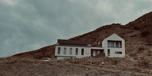 A modern house with a minimalist design is situated on a hillside. The building has a geometric, angular shape with large windows, and it is surrounded by a barren, rocky landscape. The sky is overcast, adding a moody and dramatic atmosphere.