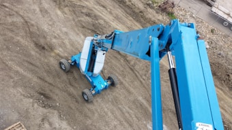 A construction vehicle with a blue articulated arm and a platform is positioned on a rough, dirt-covered surface. The vehicle has rubber tires and is situated in an area with tire tracks and scattered debris.