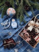 A festive arrangement with evergreen branches, a colorful Christmas ornament, a silver rocking horse, a blue matryoshka doll, and various holiday decorations placed on a blue plaid fabric. A gift box is partially visible, filled with a wrapped candy and a red and gold present ornament. Silver stars and garlands are scattered across the scene, alongside a red toy train.