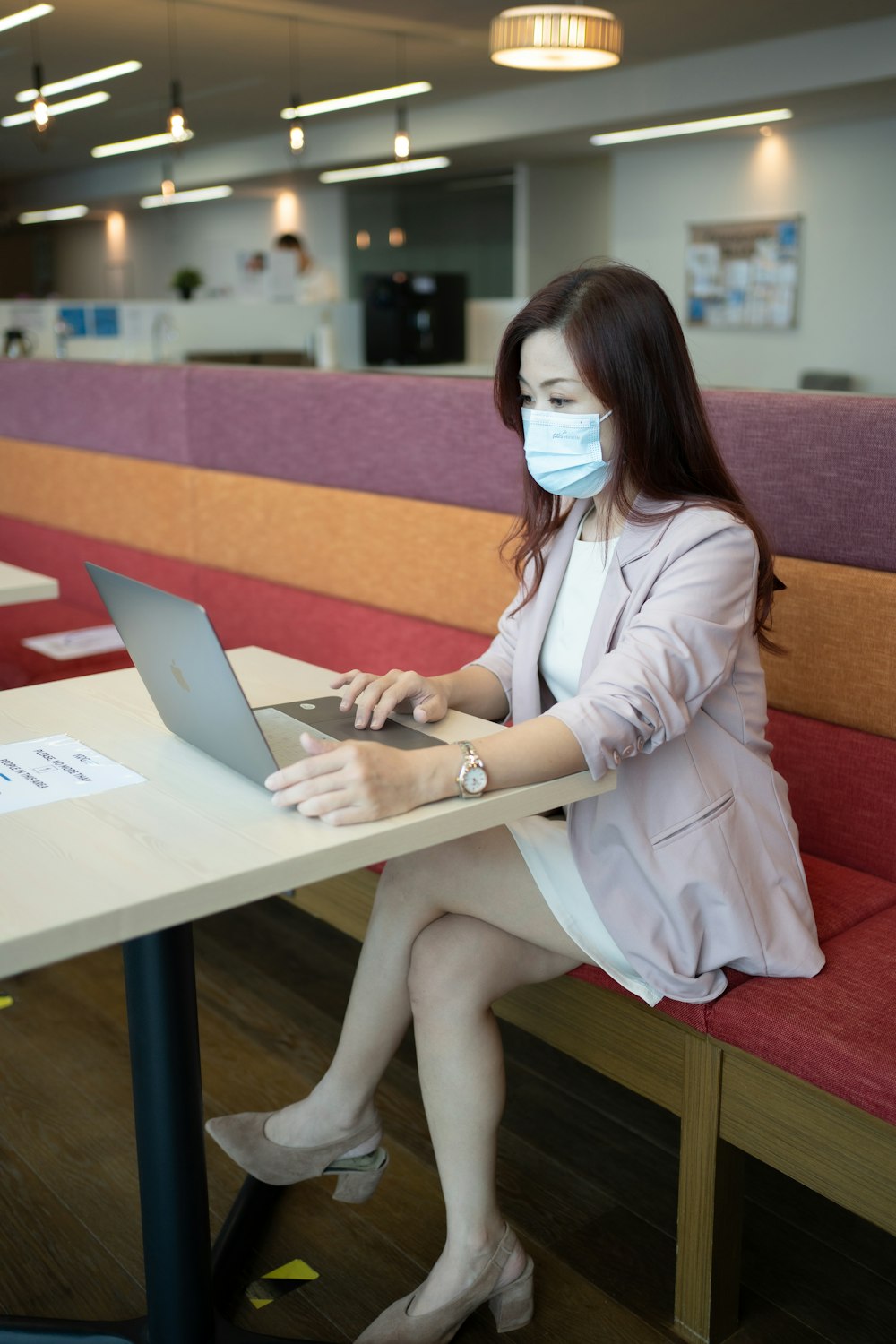 a woman sitting at a table with a laptop wearing a face mask