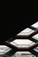 Geometric architecture with a view through several interconnected stairway platforms. The design features a series of horizontal lines and angles, framing a distant cityscape and a body of water in the background.