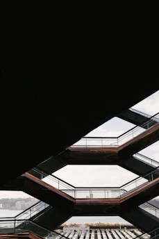 Geometric architecture with a view through several interconnected stairway platforms. The design features a series of horizontal lines and angles, framing a distant cityscape and a body of water in the background.
