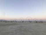 A flat, expansive desert landscape with several large circular metal structures evenly spaced across the ground. Small groups of people are gathered around these structures, indicating some event or gathering. The sky is clear with a faint gradient from pale blue to soft orange near the horizon, suggesting early morning or evening.