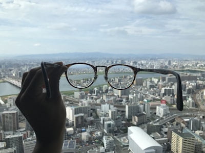 A hand holds a pair of eyeglasses against a cityscape background. The buildings are clearly seen through the lenses, while the rest of the view is slightly blurred. This contrast emphasizes the clarity and focus provided by the glasses, highlighting details of the urban landscape including skyscrapers and a river.