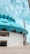The architectural structure features a modern design with a prominent, teal-colored geometric roof that resembles a flower. The building has a concrete base with a large wooden door and a spacious entrance. The sky is bright and partly cloudy, adding to the open, expansive feel of the image.