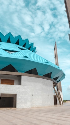 The architectural structure features a modern design with a prominent, teal-colored geometric roof that resembles a flower. The building has a concrete base with a large wooden door and a spacious entrance. The sky is bright and partly cloudy, adding to the open, expansive feel of the image.