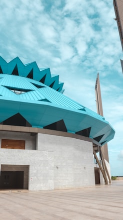 The architectural structure features a modern design with a prominent, teal-colored geometric roof that resembles a flower. The building has a concrete base with a large wooden door and a spacious entrance. The sky is bright and partly cloudy, adding to the open, expansive feel of the image.