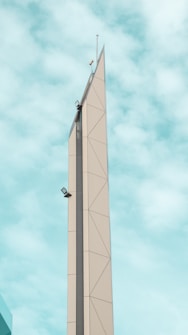 A tall, modern tower with geometric patterns ascending towards a pointed tip against a backdrop of a light blue sky with scattered clouds. The structure features a sleek design with visible seams and angles, and it is equipped with mounted equipment such as lights or sensors.