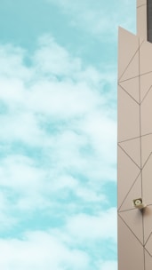 A clear view of a modern building facade featuring geometric patterns in soft beige, with a security camera mounted near the edge. The background is a bright blue sky with fluffy white clouds.