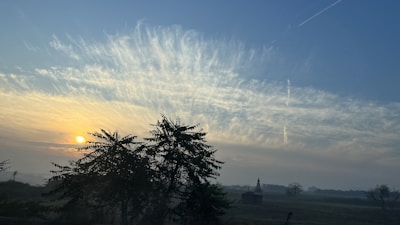 A serene landscape during sunrise with a vibrant sky displaying streaks of clouds. Silhouettes of trees and a small structure add depth to the scene. The sun emits a warm glow, casting light over the field.