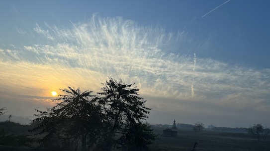 A serene landscape during sunrise with a vibrant sky displaying streaks of clouds. Silhouettes of trees and a small structure add depth to the scene. The sun emits a warm glow, casting light over the field.