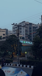 An urban street scene featuring a storefront with a glowing sign that reads 'PEYALA' in the foreground. The surrounding area includes trees, several buildings, and heavy electrical wiring overhead. There is an individual standing to the left and billboards or banners at the bottom of the image.
