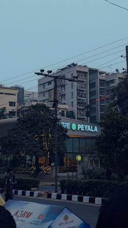 An urban street scene featuring a storefront with a glowing sign that reads 'PEYALA' in the foreground. The surrounding area includes trees, several buildings, and heavy electrical wiring overhead. There is an individual standing to the left and billboards or banners at the bottom of the image.