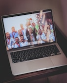 A laptop computer displaying a celebratory scene on its screen. The screen shows a group of people wearing sports uniforms and medals, enthusiastically celebrating and holding up a trophy. The keyboard is visible, and the laptop is resting on a wooden surface.