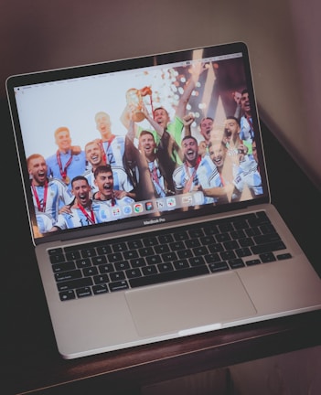 A laptop computer displaying a celebratory scene on its screen. The screen shows a group of people wearing sports uniforms and medals, enthusiastically celebrating and holding up a trophy. The keyboard is visible, and the laptop is resting on a wooden surface.