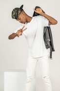 A person wearing a camouflage cap is posing playfully, holding a black jacket over one shoulder and flashing a peace sign with one hand. Dressed in a white t-shirt and white pants, the individual stands against a plain background.
