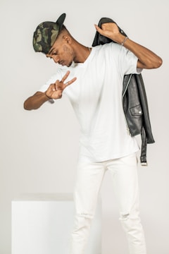 A person wearing a camouflage cap is posing playfully, holding a black jacket over one shoulder and flashing a peace sign with one hand. Dressed in a white t-shirt and white pants, the individual stands against a plain background.