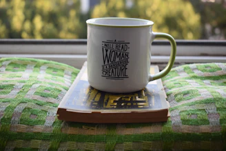 A white ceramic mug with green trim sits on top of a closed book. The mug has a printed quote that reads, 'Well-read woman is a dangerous creature'. The book and mug are placed on a green patterned fabric, with a blurred view of trees outside a window in the background.