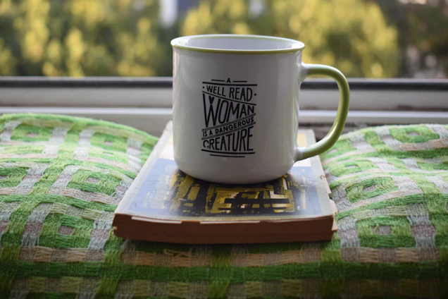 A white ceramic mug with green trim sits on top of a closed book. The mug has a printed quote that reads, 'Well-read woman is a dangerous creature'. The book and mug are placed on a green patterned fabric, with a blurred view of trees outside a window in the background.