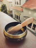 A Tibetan singing bowl with decorative patterns and a wooden mallet resting inside, positioned on a wooden ledge. In the background, several houses with light blue and beige walls and tiled roofs are visible, blurred out of focus.