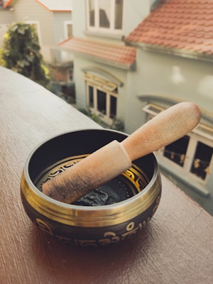 A Tibetan singing bowl with decorative patterns and a wooden mallet resting inside, positioned on a wooden ledge. In the background, several houses with light blue and beige walls and tiled roofs are visible, blurred out of focus.
