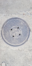 A metal manhole cover embedded in the concrete ground, featuring a grid pattern and several small holes. The surface around the cover shows rough, textured concrete with small stones.