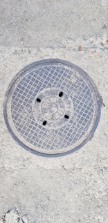 A metal manhole cover embedded in the concrete ground, featuring a grid pattern and several small holes. The surface around the cover shows rough, textured concrete with small stones.