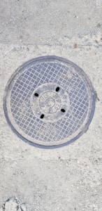 A metal manhole cover embedded in the concrete ground, featuring a grid pattern and several small holes. The surface around the cover shows rough, textured concrete with small stones.