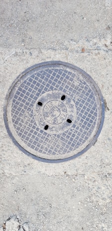 A metal manhole cover embedded in the concrete ground, featuring a grid pattern and several small holes. The surface around the cover shows rough, textured concrete with small stones.